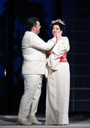 Vergrößerte Ansicht von Ein Mann in hellem Anzug berührt sanft den Hals einer Frau in weißem Kimono mit rotem Obi und Blumenkranz im Haar vor dunklem Hintergrund