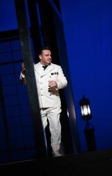 Vergrößerte Ansicht von Mann in weißem Anzug mit schwarzen Knöpfen und Abzeichen steht hinter schwarzem Metallrahmen vor blauem Hintergrund mit Laterne