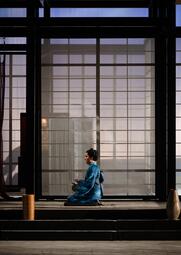 Vergrößerte Ansicht von Person in traditionellem blauen Kimono sitzt auf dem Boden vor einem großen Fenster mit Gitterstruktur