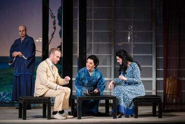 Vergrößerte Ansicht von Drei Personen sitzen an einem niedrigen Tisch, eine steht im Hintergrund, alle tragen unterschiedliche Kleidung, darunter ein blauer Kimono und ein helles Anzug-Outfit