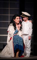 Vergrößerte Ansicht von Eine Frau in einem blauen, gemusterten Kleid und weißem Umhang kniet neben einem Jungen in weißer Matrosenuniform mit Mütze vor einem dunklen Hintergrund