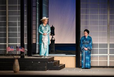 Vergrößerte Ansicht von Zwei Personen in traditioneller und historischer Kleidung stehen auf einer Bühne mit japanisch anmutenden Schiebetüren und amerikanischen Flaggen in einem Gefäß.