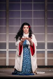 Vergrößerte Ansicht von Eine Frau kniet in einem blauen, gemusterten Kleid mit weißem Umhang und hält eine Schale mit einem Schmetterling darin