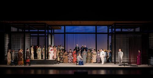 Vergrößerte Ansicht von Bühnenbild mit einer großen Gruppe von Personen in traditioneller japanischer Kleidung und westlicher Kleidung, die auf einer Bühne mit japanisch inspiriertem Hintergrund stehen