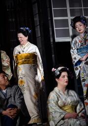 Vergrößerte Ansicht von Mehrere Personen in traditionellen japanischen Kimonos mit floralen Mustern, zwei Frauen stehen, eine sitzt, alle mit Haarschmuck, im Hintergrund ein Fenster mit Gittern.
