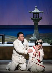 Vergrößerte Ansicht von Ein Mann in weißem Anzug kniet neben einer Frau in traditionellem japanischem Kimono mit Blumenhaarschmuck vor einer steinernen Laterne auf einer Bühne.