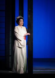 Vergrößerte Ansicht von Person in traditionellem hellen Kimono mit Blumenkranz auf dem Kopf steht an einem schwarzen Pfosten vor blauem Hintergrund