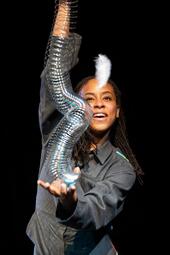 Vergrößerte Ansicht von Frontalaufnahme von einer Frau in einer grauen Jacke, die auf der einen Handfläche eine Spirale in der Hand hält, die sie mit der anderen Hand hochhält. Vor ihr fliegt eine Vogelfeder in der Luft.