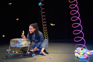 Vergrößerte Ansicht von Eine Frau sitzt auf dem Boden vor einem Ventilator und um sie herum liegen bunte Federn und ein Ventilator. Fasziniert hebt sie Federn über den Ventilator.Bunte Federn fliegen durch die Luft und bunte Spiralen liegen auf dem Boden.