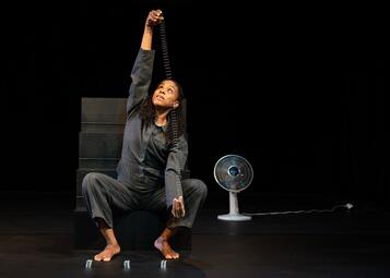 Vergrößerte Ansicht von Eine Frau geht in die Hocke und hebt eine Metallspirale mit beiden Händen fest und zieht sie auseinander. Sie schaut nach oben zu ihrer einen Hand. Vor ihr stehen drei weitere silberne Spiralen auf dem Boden. Im Hintergrund steht ein Ventilator.