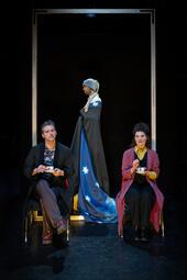 Vergrößerte Ansicht von Drei Personen sind zu sehen. Zwei Menschen sitzen auf Stühlen mit Teetassen in den Händen. Die dritte Person trägt einen Turban, ein langes, blaues Gewand mit Sternen und läuft hinter den Stühlen nach links.