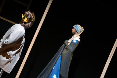 Vergrößerte Ansicht von Zwei Personen. Die Linke ist mit dem Rücken zu uns gedreht. Sie trägt einen weißen Mantel und eine goldene Krone. Die rechte Person ist im Seitenprofil zu sehen. Er trägt einen Turban und einen schwarzen Umhang über ein dunkelblaues Sternengewand.