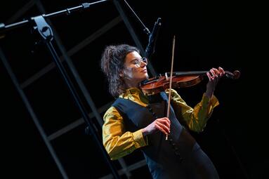 Vergrößerte Ansicht von Eine Person, die Geige spielt. Sie trägt eine schwarze Hose und Weste über ein gelbes Hemd.