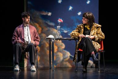 Vergrößerte Ansicht von Zwei Personen sitzen an einem Tisch. Die Linke trägt einen alt-rosa Mantel über ein weißes Hemd und schwarze Hosen. Die zweite Person trägt eine Brille und lange, braune Haare. Sie trägt einen leopardengemusterten Pelzmantel über einen schwarzen…