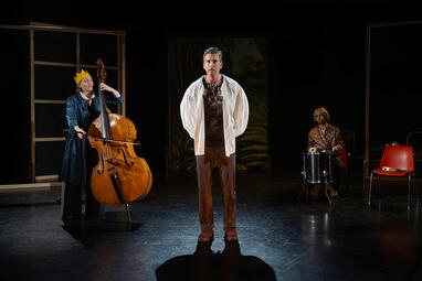 Vergrößerte Ansicht von Drei Menschen sind in einer frontalen Einstellungen zu sehen. Die Person in der Mitte trägt ein weißes Hemd über ein gemustertes Shirt und braune Hosen. Die Person zu seiner linken spielt Cello und trägt eine goldene Papierkrone und einen langen,…