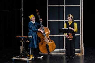 Vergrößerte Ansicht von Zwei Personen sind zu sehen. Die linke Person steht hinter einem Keyboard und hält ein Cello. Sie trägt ein langes, dunkelblaues Gewand und eine goldene Papierkrone. Die rechte Person trägt eine goldene Krone, eine schwarze Hose und eine schwarze…
