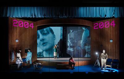 Vergrößerte Ansicht von Mehrere Personen in einer Bühnenkulisse mit einem hölzernen, schräg stehenden Raum und einem Mikrofonständer im Vordergrund