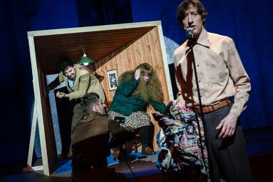 Vergrößerte Ansicht von Bühnenbild mit Holzwand, blauer Bühne und blauen Vorhängen, Projektion von zwei Personen, zwei Mal die Zahl '2004' in Pink, fünf Personen auf der Bühne in verschiedenen Posen und Kleidung, darunter eine Person vor einer Kamera mit Lichtquelle.