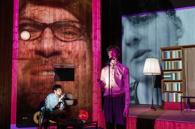 Vergrößerte Ansicht von Person steht mit Mikrofon in einem Raum mit Bücherregal, Stehlampe und großem schwarz-weißem Gesichtsporträt an der Wand