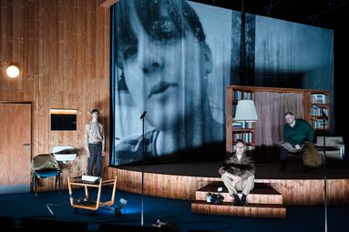 Vergrößerte Ansicht von Bühnenbild mit Holzpaneelwand, zwei Personen, einem Stuhl, einem kleinen Tisch und einem Bücherregal mit Lampe