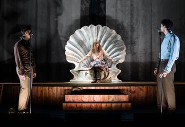 Vergrößerte Ansicht von Person sitzt auf einer großen Muschel-förmigen Bühne, zwei Personen stehen jeweils links und rechts davon mit Mikrofonen