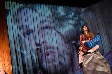 Vergrößerte Ansicht von Person in historischer Kleidung sitzt auf einem Felsen und hält einen blauen Wäschekorb vor einer Holzwand mit großem projiziertem Gesicht im Hintergrund