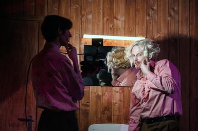 Vergrößerte Ansicht von Person mit hochgestecktem Haar und rosa Hemd filmt eine weitere Person mit lockigem Haar und rosa Hemd vor einem Spiegel in einem holzvertäfelten Raum