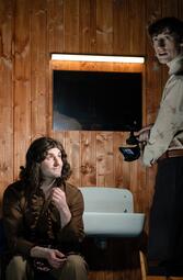 Vergrößerte Ansicht von Zwei Personen in einem Raum mit holzvertäfelter Wand, eine sitzt vor einem weißen Waschbecken, die andere steht und hält eine Kamera
