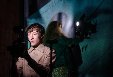 Vergrößerte Ansicht von Person mit hellblauem Hemd bedient eine Kamera vor einer Wandprojektion mit unscharfer Person und Text