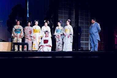 Vergrößerte Ansicht von Gruppe von sieben Personen in traditionellen japanischen Kimonos und ein Mann in einem hellen Anzug auf einer Bühne mit dunklem Hintergrund