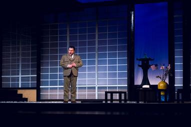 Vergrößerte Ansicht von Mann in braunem Anzug steht auf Bühne vor Gitterwand mit japanischer Statue, Vase und US-Flaggen