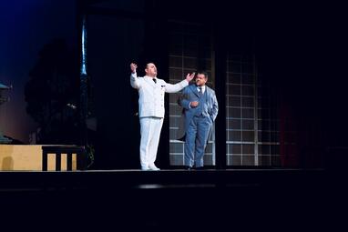 Vergrößerte Ansicht von Zwei Männer auf einer Bühne, einer in weißem Anzug mit erhobenen Armen, der andere in grauem Anzug mit Hand in der Hosentasche