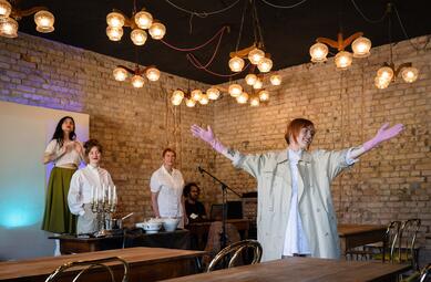 Vergrößerte Ansicht von Sari Dorian steht mit breit erhobenen Armen im Raum und schaut lächelnd nach rechts. Sie trägt rosa Gummihandschuhe. Maria Stickel und Claudia Pflaum-Richter schauen ihr leicht lächelnd zu, Yasmin Ahmed hält ihre Hände über eine imaginäre Kuppel und…