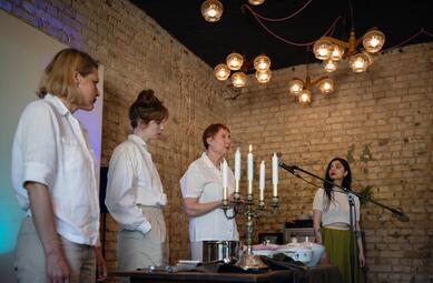 Vergrößerte Ansicht von Schräge Ansicht. Tina Stottko (links) und Yasmin Ahmed (rechts) stehen am Tisch und schauen Claudia Plaum-Richter an, während sie ins Standmikrofon spricht. Maria Stickel schaut mit ihren händen in den Hosentaschen in den Kochtopf.