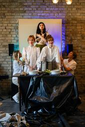 Vergrößerte Ansicht von Sari Dorian und Maria Stickel stehen vor dem Tisch und schauen leicht lächelnd durch den Raum. rechts und links von ihnen knien Tina Stottko und Claudia Pflaum-Richter, welche einen weißen rahmen hinter Yasmin Ahmed halten. Ahmed steht erhöht und…