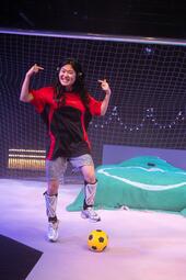 Vergrößerte Ansicht von Eine Person steht vor einem Fußballtor und trägt ein rot-schwarzes Trikot, silberfarbene Shorts und silberne Schuhe. Die Beine sind mit Schienbeinschonern geschützt. Ein gelber Fußball liegt vor dem Fuß. Im Hintergrund ist ein Netz mit Lichterketten…