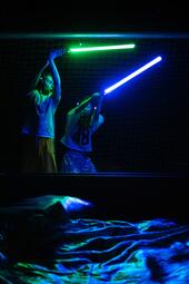 Vergrößerte Ansicht von Zwei Personen stehen nebeneinander und halten jeweils einen leuchtenden Stab, einer grün, einer blau. Die Szene ist dunkel, die Lichtstäbe werfen farbige Reflexionen auf die Umgebung. Im Vordergrund liegt ein Stoff oder Kleidungsstück, das ebenfalls…