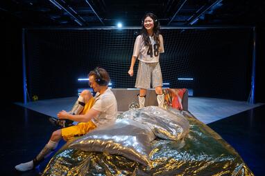 Vergrößerte Ansicht von Zwei Personen befinden sich auf einer Bühne vor einem Fußballtor. Eine Person sitzt mit angewinkelten Beinen auf einer glänzenden, silberfarbenen Decke und trägt gelbe Shorts, ein weißes Trikot sowie Kopfhörer. Die andere Person steht auf der Decke…