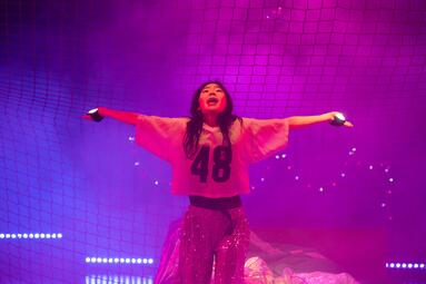 Vergrößerte Ansicht von Eine Person steht mit ausgebreiteten Armen vor einem Netz im Hintergrund. Sie trägt ein helles Trikot mit der Nummer „48“ und eine glitzernde Hose. Die Bühne ist in pink-violettes Licht getaucht, hinter der Person sind Lichterketten und ein grünes…