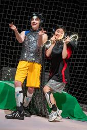 Vergrößerte Ansicht von Zwei Personen stehen nebeneinander vor einem Netz auf der Bühne. Beide tragen Kopfhörer und sportliche Kleidung mit glänzenden Elementen. Eine Person trägt gelbe Shorts und ein silbernes Oberteil mit Umhang, die andere ein rot-schwarzes Trikot mit…