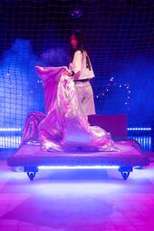 Vergrößerte Ansicht von Eine Person steht auf einem niedrigen, rollbaren Bett und hält eine große, silberglänzende Decke in den Händen. Sie trägt ein weißes Trikot mit der Nummer „48“ und silberfarbene Shorts. Das Bühnenbild besteht aus einem großflächigen Netz im…