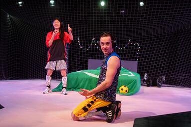 Vergrößerte Ansicht von Eine Person kniet im Vordergrund auf der Bühne und trägt ein silbernes Oberteil, gelbe Shorts und Schienbeinschoner. Im Hintergrund steht eine zweite Person vor einem Netz mit Lichterketten. Zwischen ihnen befindet sich ein Bett, das mit einer…