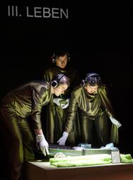 Vergrößerte Ansicht von Drei Personen in glänzenden Anzügen und weißen Handschuhen beugen sich über einen beleuchteten Tisch mit einem Modell und tragen Kopfhörer