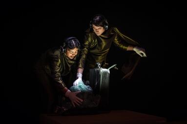 Vergrößerte Ansicht von Zwei Personen in glänzenden Anzügen und weißen Handschuhen greifen nach einem mit Luftpolsterfolie umwickelten Gegenstand neben einem silbernen Koffer in dunkler Umgebung