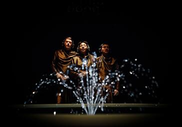 Vergrößerte Ansicht von Drei Personen in dunkler Kleidung stehen hinter einem beleuchteten Springbrunnen vor schwarzem Hintergrund