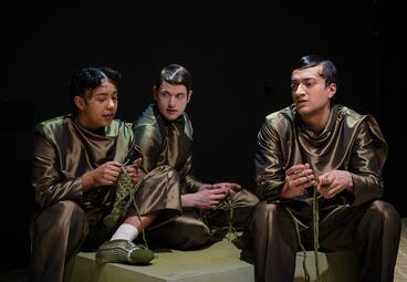 Vergrößerte Ansicht von Drei Personen in glänzenden, einfarbigen Outfits sitzen auf einer Bühne und stricken mit grüner Wolle.