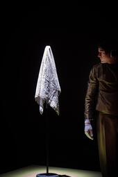 Vergrößerte Ansicht von Ein weißes Tuch hängt auf einem Ständer vor schwarzem Hintergrund, rechts daneben steht eine Person in brauner Kleidung und weißen Handschuhen.
