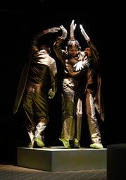 Vergrößerte Ansicht von Drei Personen in glänzenden Anzügen und weißen Handschuhen stehen auf einem Podest und heben die Arme in die Luft