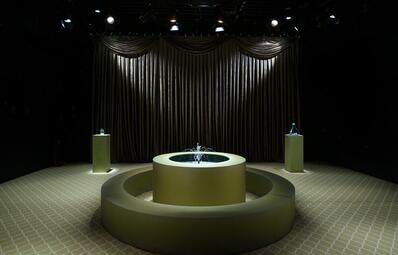 Vergrößerte Ansicht von Runder Springbrunnen mit Wasserstrahlen in einem Raum mit dunklen Vorhängen und zwei beleuchteten Sockeln