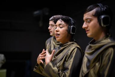 Vergrößerte Ansicht von Drei Personen mit Kopfhörern und braunen Umhängen in einer dunklen Umgebung, eine Person gestikuliert mit den Händen